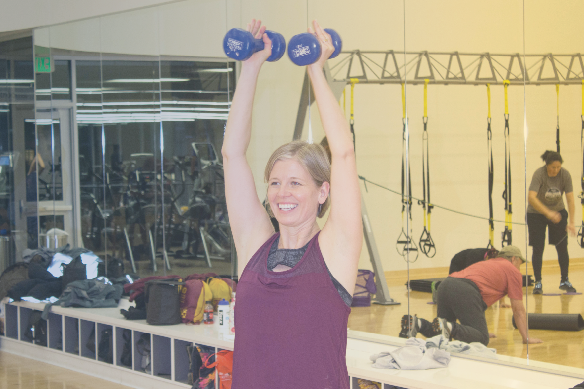 instructor lifting two weights above head while leading the class